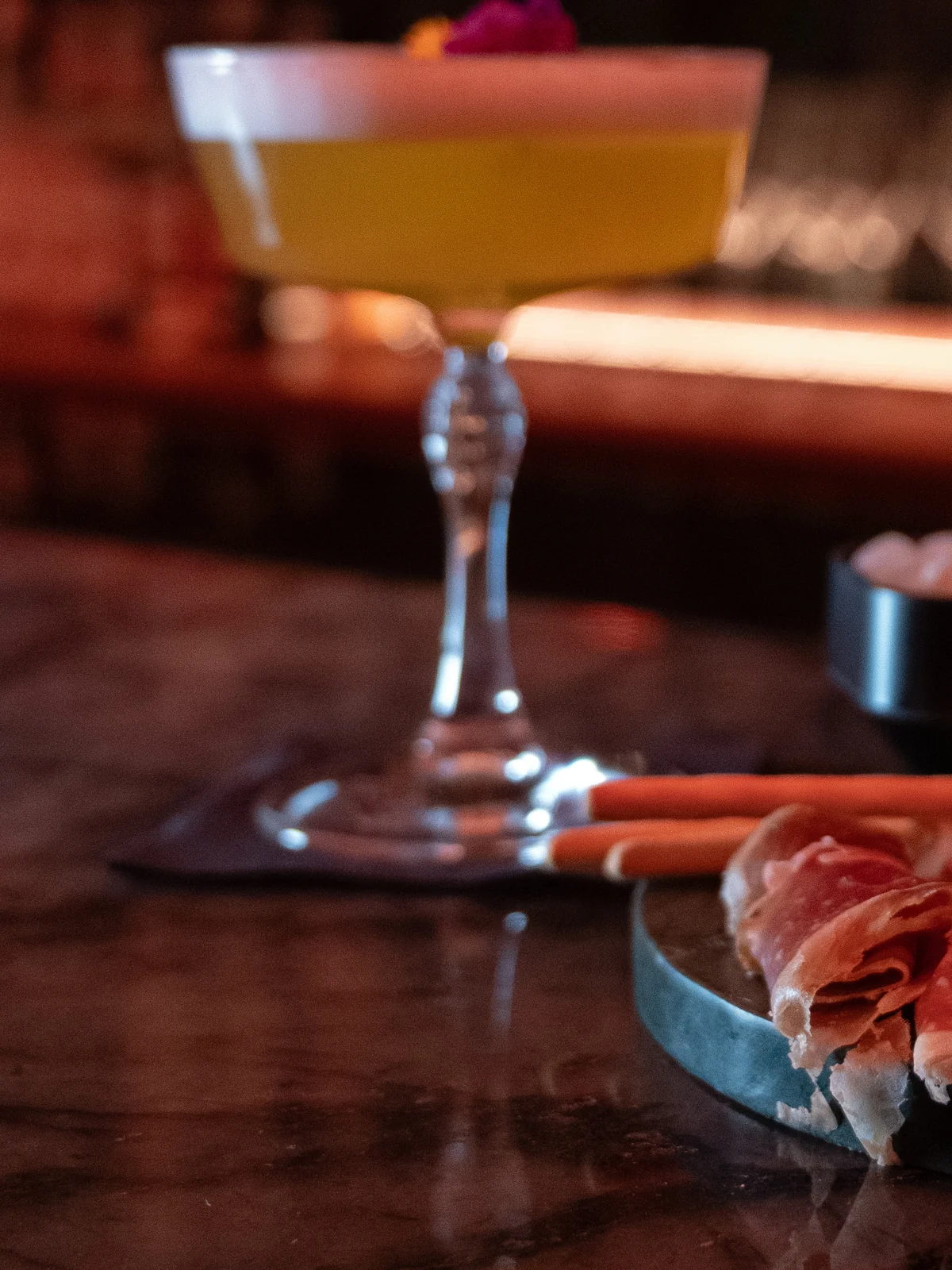A cocktail in a coupe next to a plate of ham on the bar at Mayfair